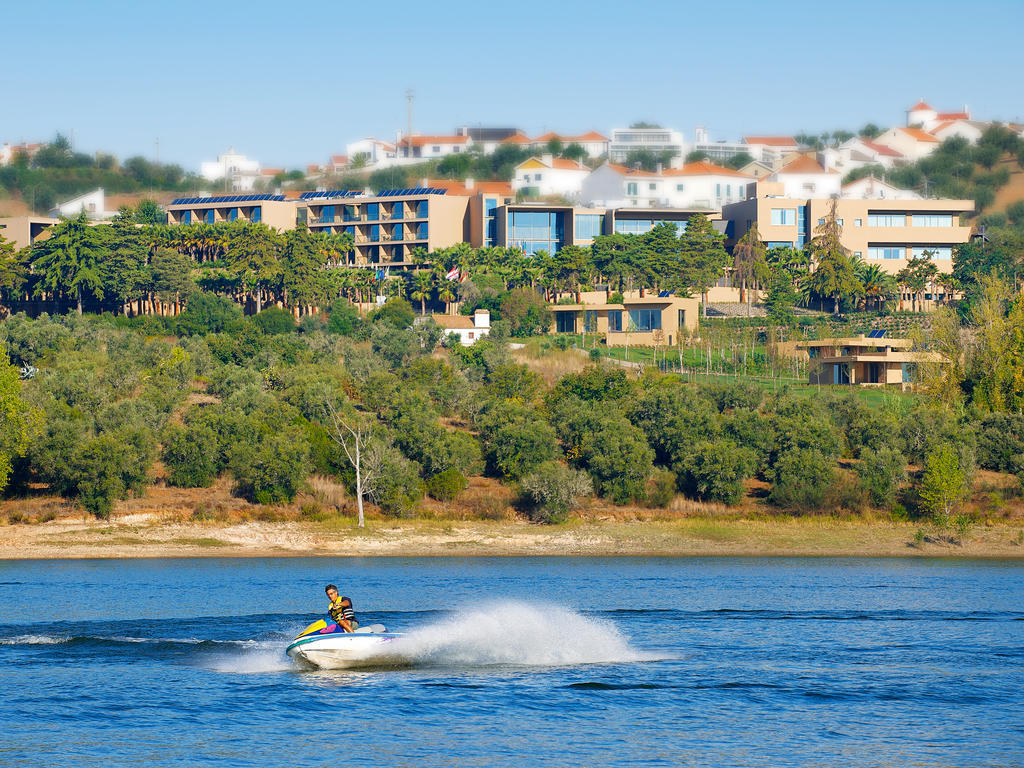 Ap Lago Montargil - Conference & Spa Екстериор снимка The photo depicts a person riding a jet ski on a body of water. In the background, there are modern buildings and lush greenery, suggesting a scenic landscape, likely near a resort or upscale residential area. The clear blue sky and calm water enhanc