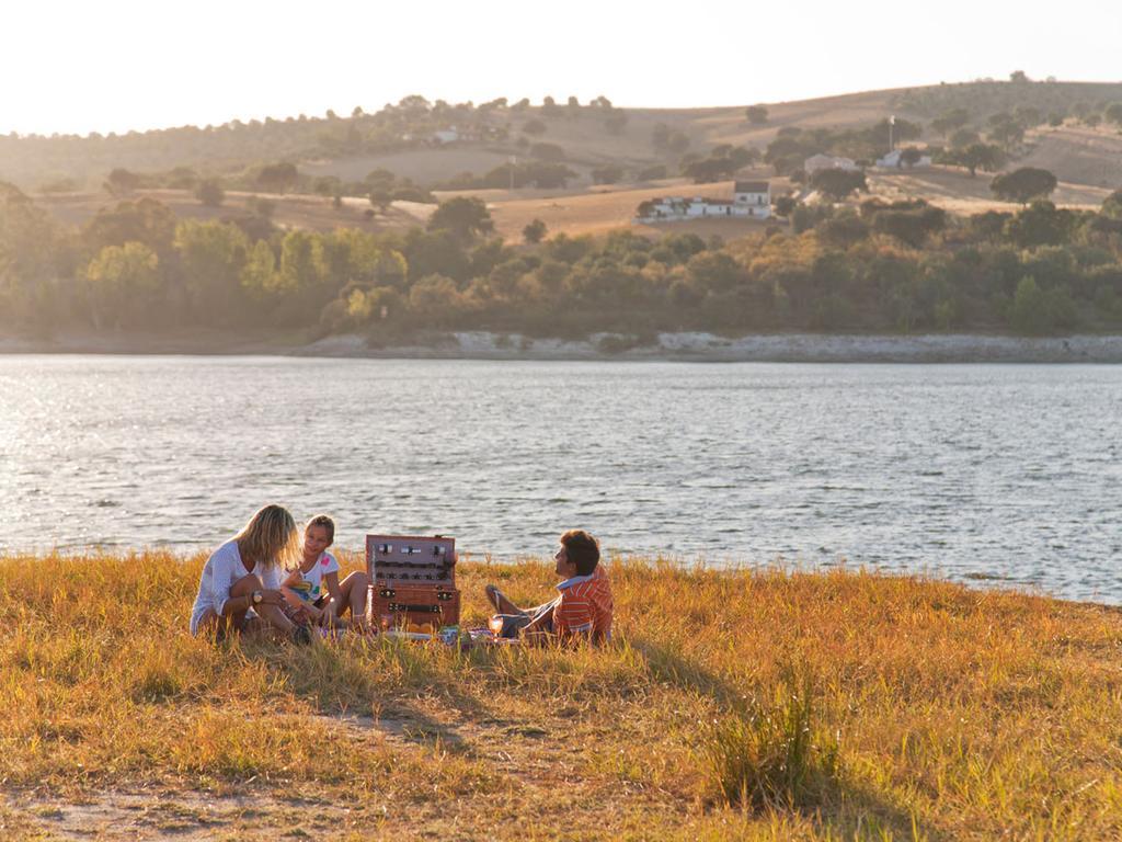 Ap Lago Montargil - Conference & Spa Екстериор снимка The photo depicts a serene lakeside scene during sunset. Three people are gathered on a grassy area near the water, enjoying a picnic. They are seated around a picnic basket, with the shimmering lake in the background and hills in the distance. The w