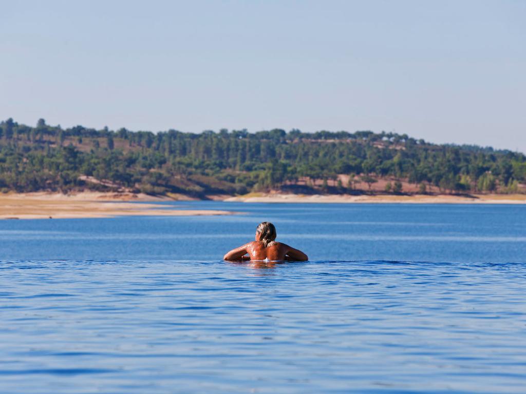 Ap Lago Montargil - Conference & Spa Екстериор снимка The photo shows a person swimming in a calm body of water, likely a lake or a river. The swimmer is seen from behind and appears to be enjoying the water on a sunny day. In the background, there's a scenic landscape featuring green trees and a sandy 