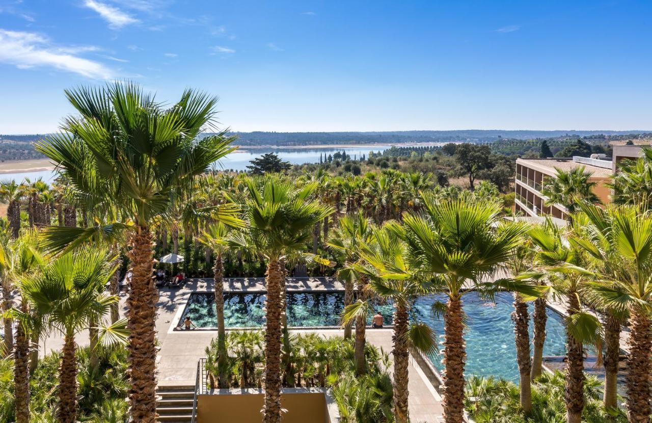 Ap Lago Montargil - Conference & Spa Екстериор снимка The photo depicts a picturesque resort landscape with a clear blue sky. In the foreground, there are tall palm trees surrounding a swimming pool area. The swimming pool has a modern design and is surrounded by lush greenery. In the background, you ca