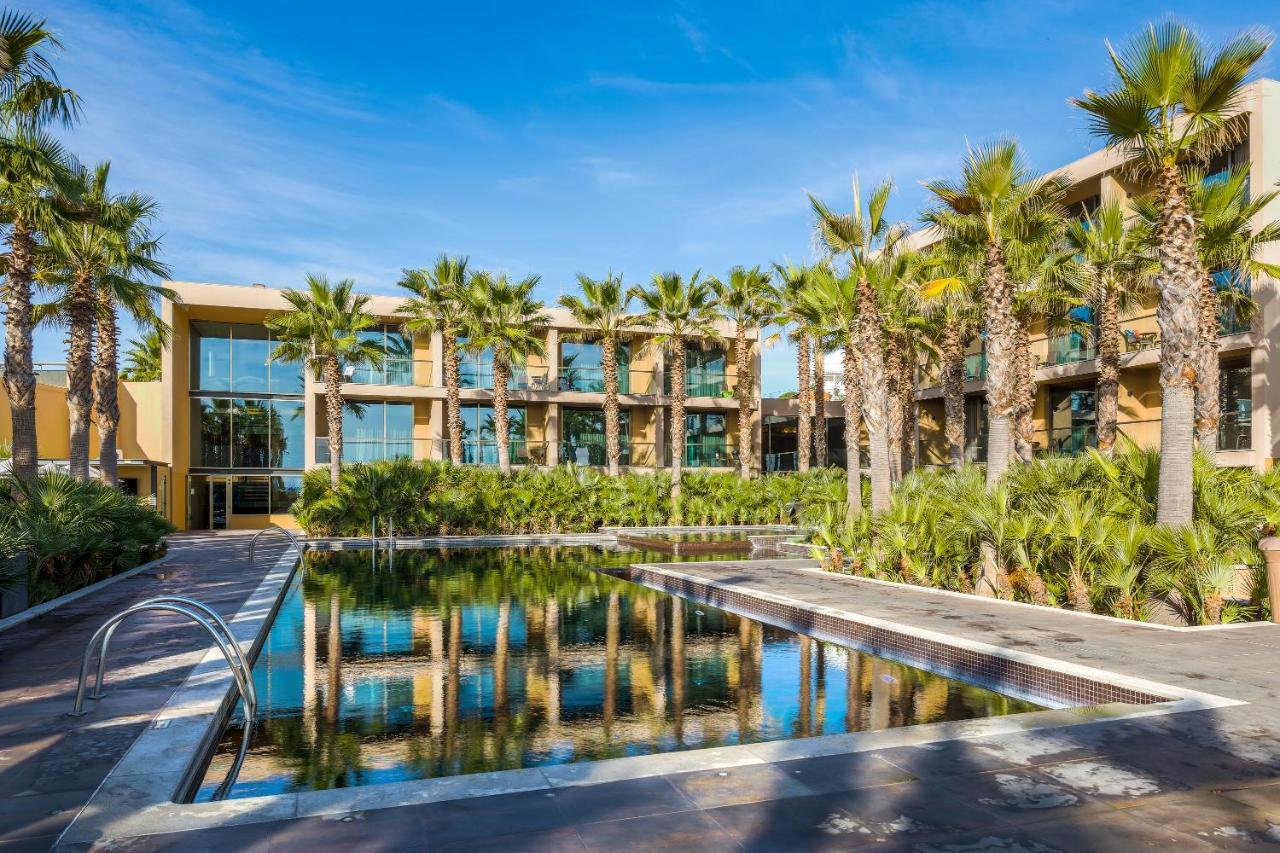 Ap Lago Montargil - Conference & Spa Екстериор снимка The photo features a modern building surrounded by lush greenery and palm trees. In the foreground, there is a serene swimming pool with a reflective surface that captures the surrounding environment. The architecture of the building appears to have 