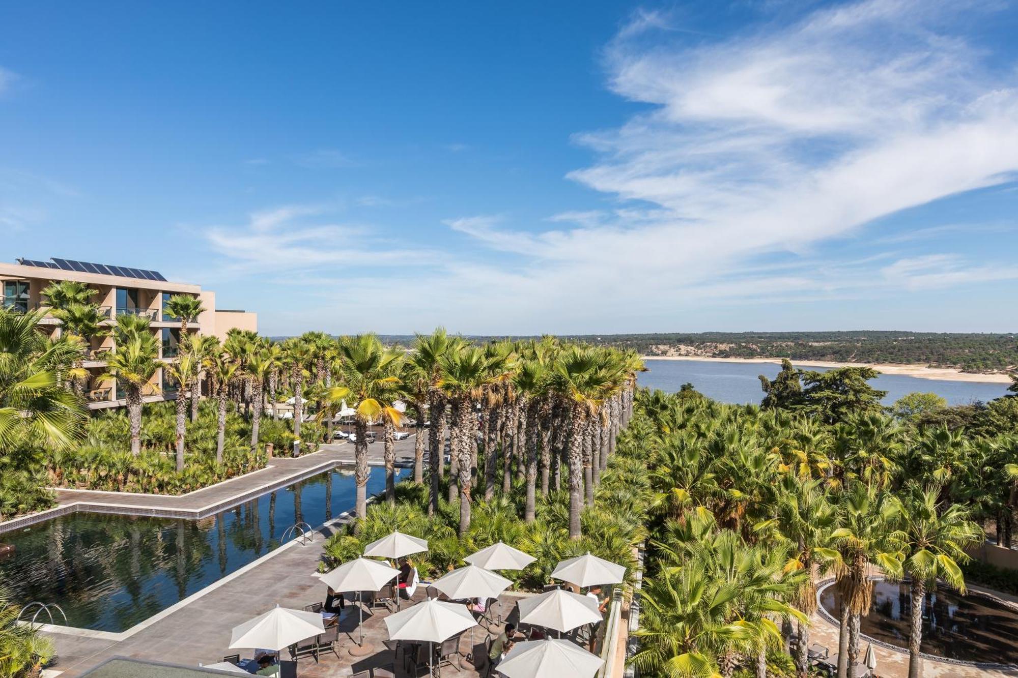 Ap Lago Montargil - Conference & Spa Екстериор снимка The photo shows a scenic view of a resort or hotel area. In the foreground, there is a pool surrounded by palm trees and lounge chairs with umbrellas, suggesting a relaxing atmosphere. Beyond the pool, there is a body of water, likely a river or coas