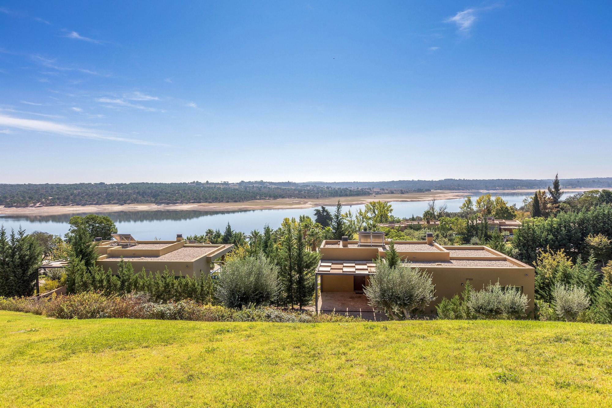 Ap Lago Montargil - Conference & Spa Екстериор снимка The photo depicts a scenic landscape featuring a calm body of water, possibly a lake or river, surrounded by lush greenery and trees. In the foreground, there are several modern buildings with flat roofs, set against a vibrant blue sky. The overall s