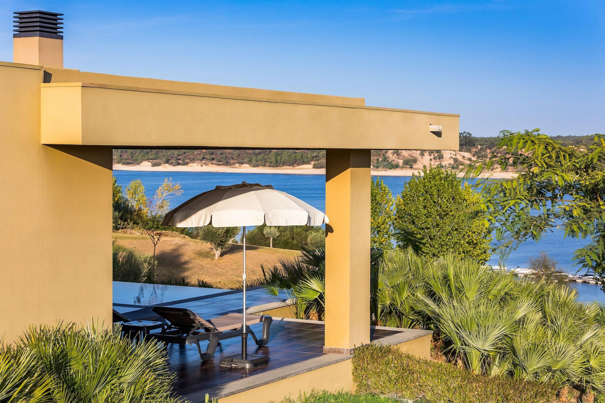 Ap Lago Montargil - Conference & Spa Екстериор снимка The photo shows a peaceful outdoor area with a modern architectural structure featuring an overhang. There is a lounge chair positioned beneath a large umbrella, creating a shaded spot to relax. In the background, there is a picturesque view of a bod