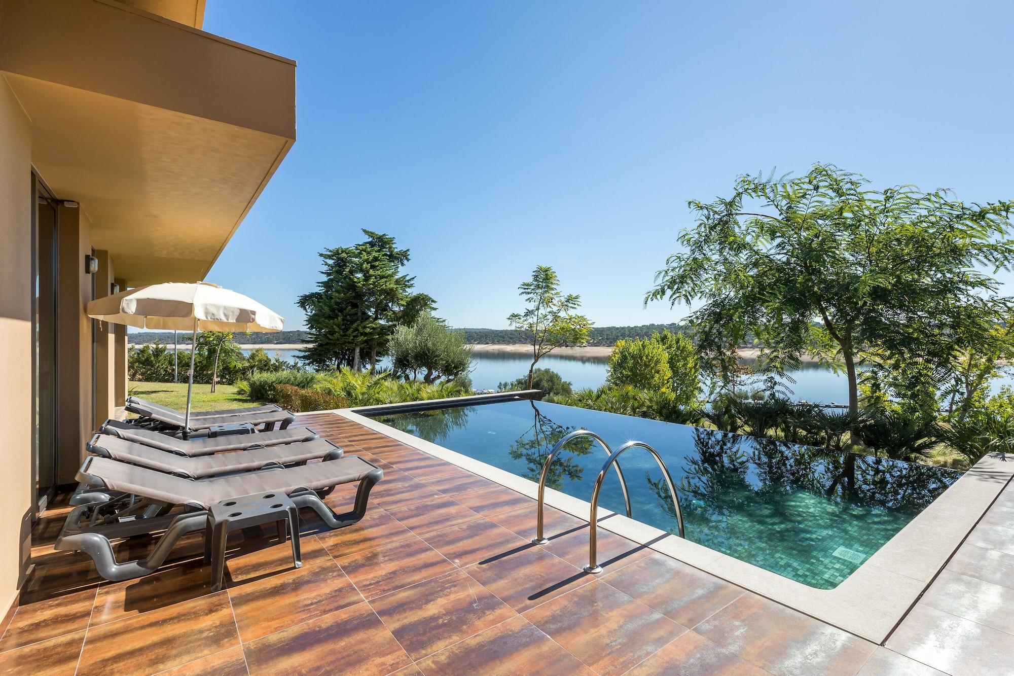 Ap Lago Montargil - Conference & Spa Екстериор снимка The photo shows a luxurious outdoor setting by a pool. There are several lounge chairs arranged next to an infinity pool, which overlooks a scenic body of water and lush greenery. A parasol provides shade for relaxation. The sky is clear and blue, in