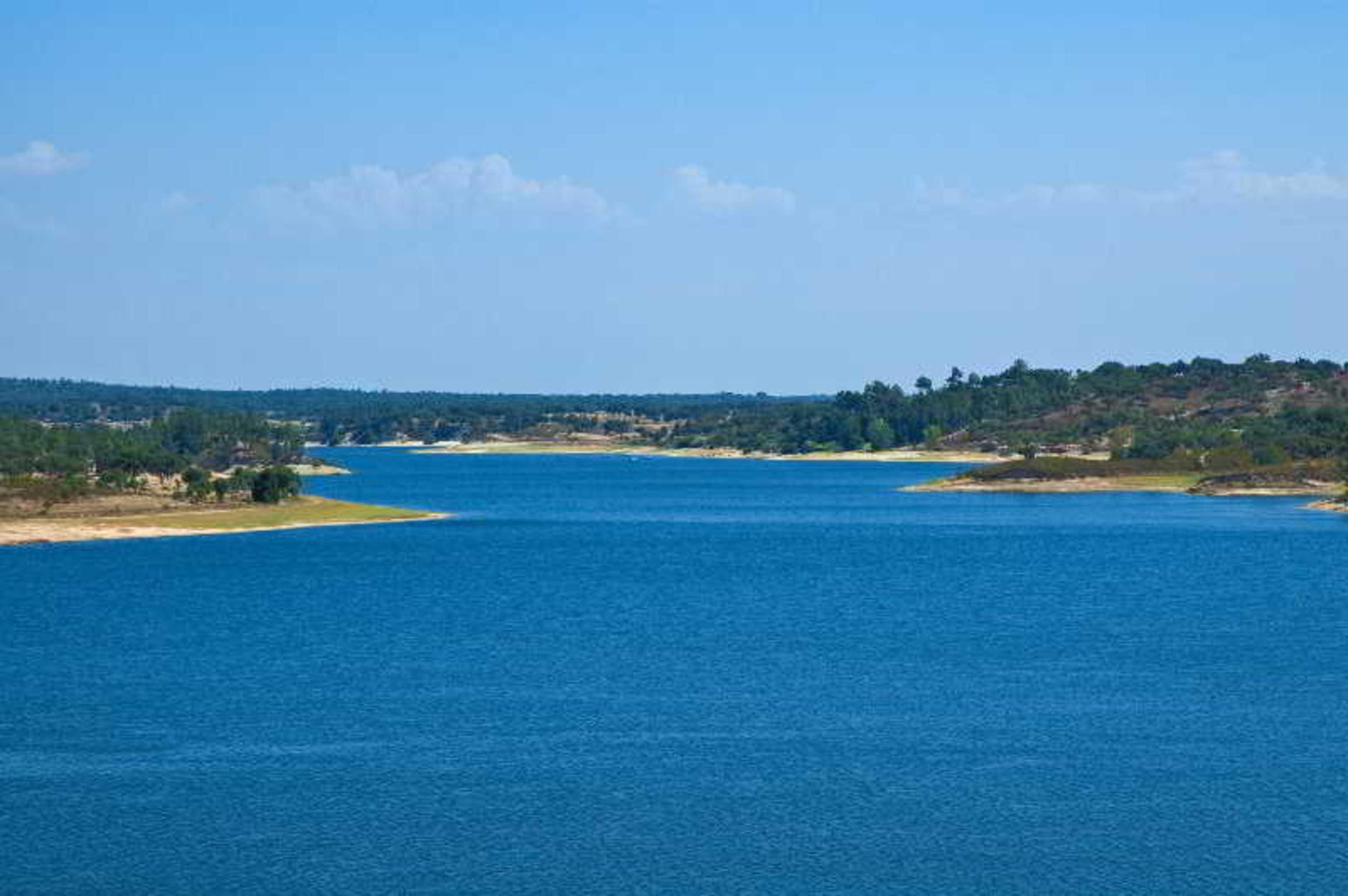 Ap Lago Montargil - Conference & Spa Екстериор снимка The photo depicts a serene landscape featuring a large body of water, likely a lake or river. The water appears calm and is reflecting a clear blue sky with some clouds. Surrounding the water, there are patches of green land and trees, indicating a n
