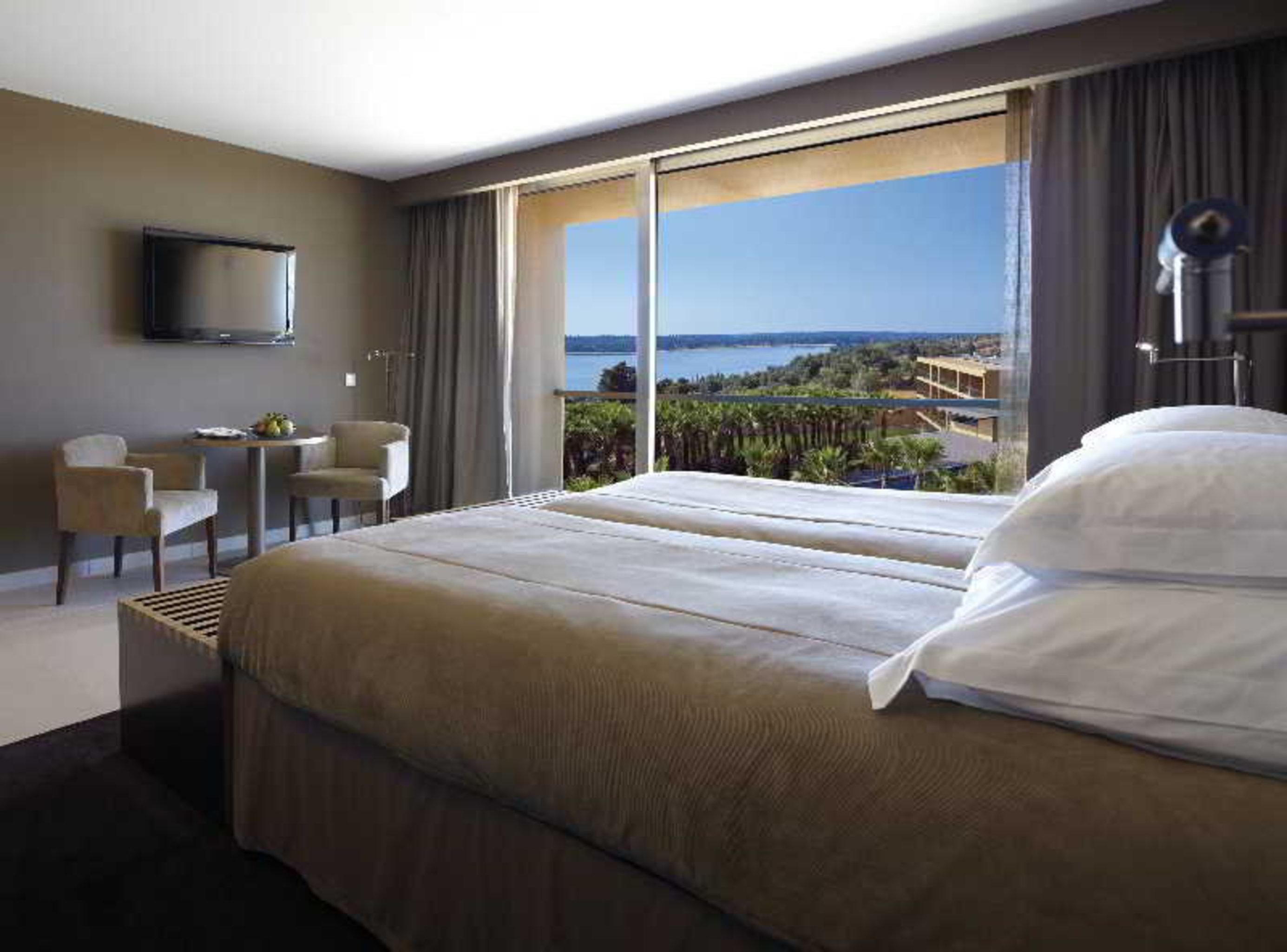 Ap Lago Montargil - Conference & Spa Екстериор снимка The photo shows a modern hotel room with a large window offering a view of the sea and mountains. The room features a comfortable bed with white pillows and brown bedding. There is a small sitting area with two chairs and a table, as well as a wall-m
