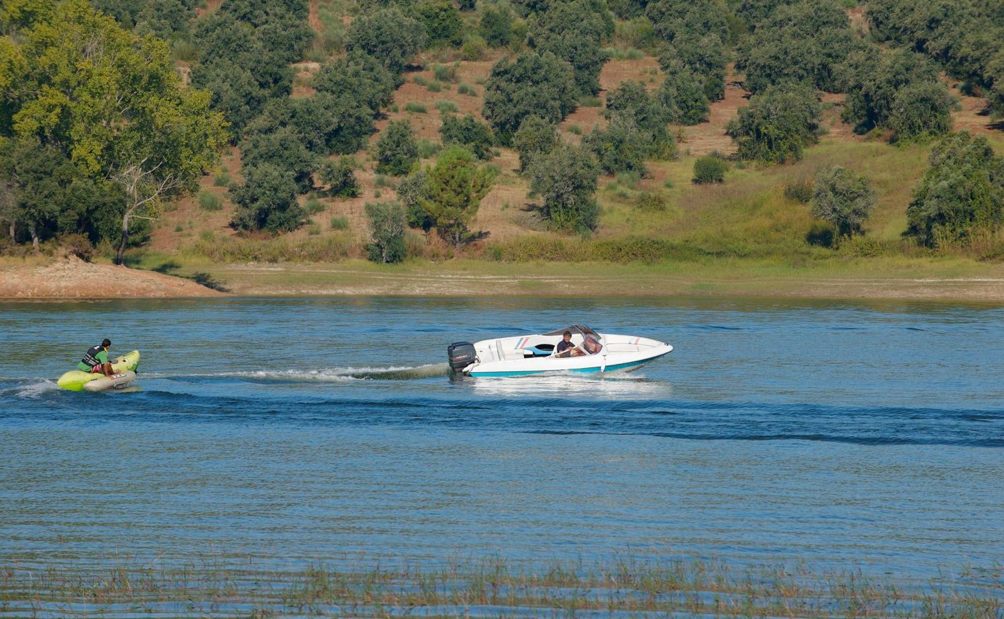 Ap Lago Montargil - Conference & Spa Екстериор снимка The photo shows a scene on a water body, likely a river or lake. In the foreground, a speedboat is moving quickly through the water, leaving a wake behind it. There are two people visible in the boat. In the background, there is a hilly area covered 