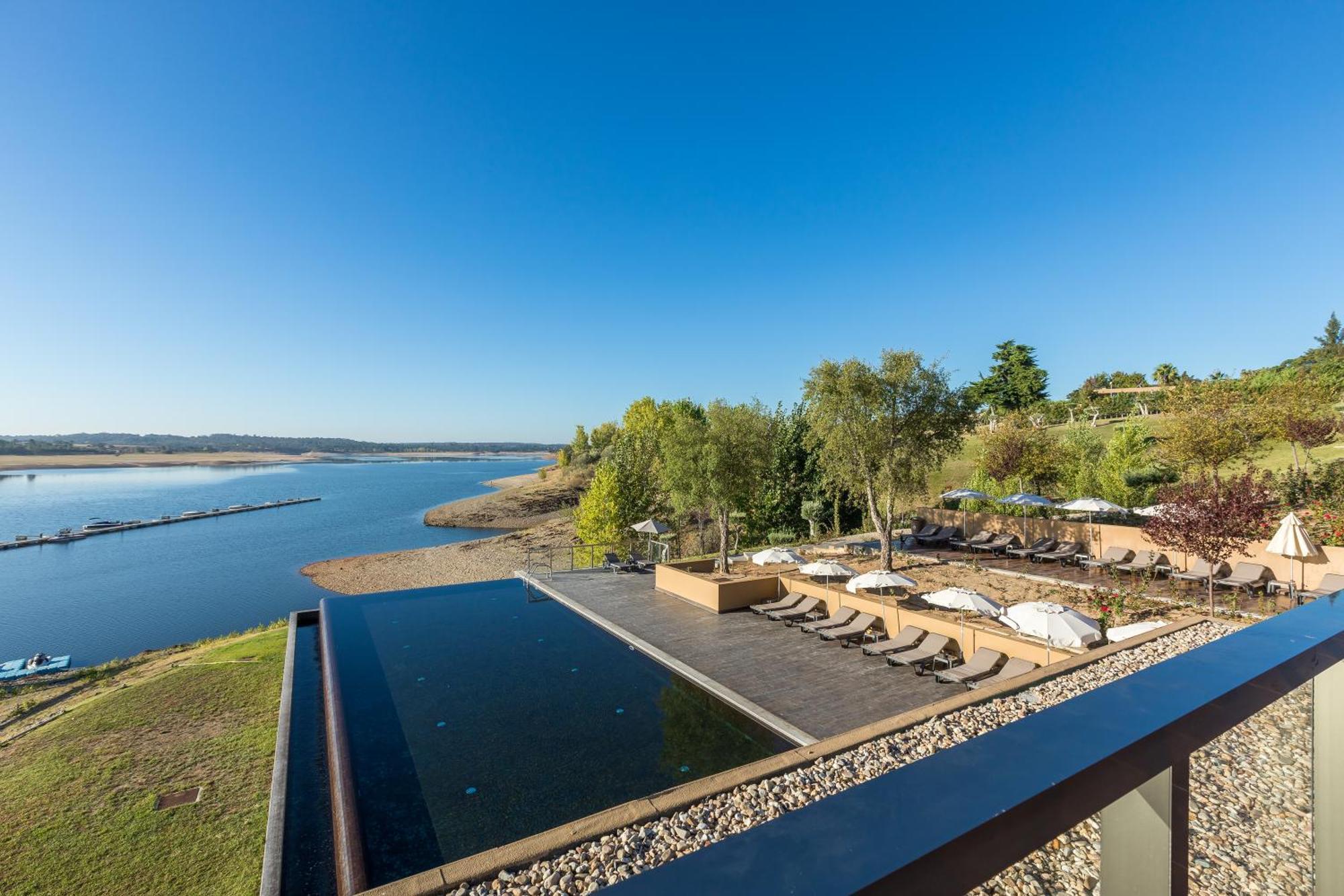 Ap Lago Montargil - Conference & Spa Екстериор снимка The photograph depicts a scenic view of a tranquil water body, likely a lake or river, bordered by lush greenery. In the foreground, there is an infinity pool that blends seamlessly with the water beyond. Comfortable lounge chairs are arranged on a w
