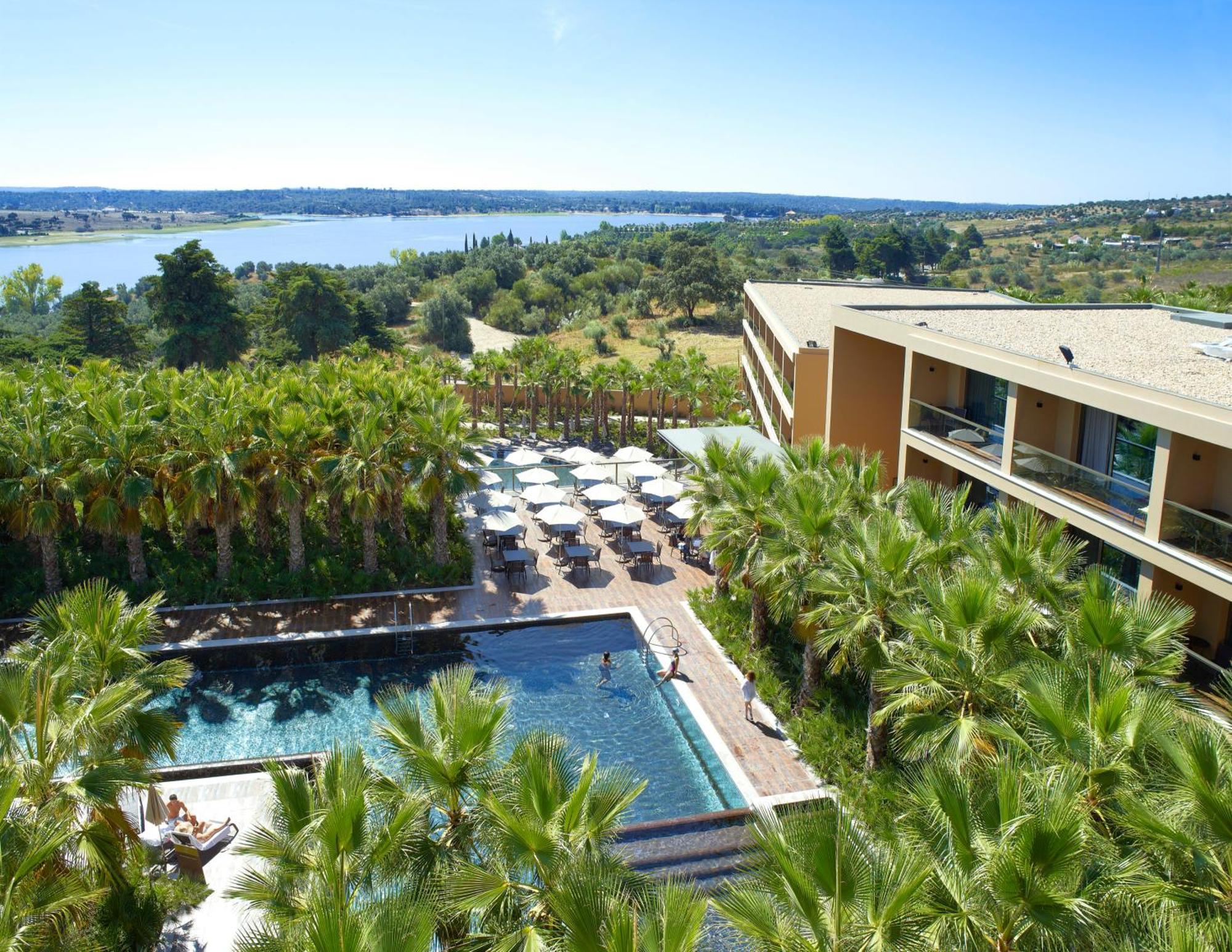 Ap Lago Montargil - Conference & Spa Екстериор снимка The photo shows a scenic view of a modern hotel or resort. In the foreground, there is a swimming pool surrounded by palm trees, with lounge chairs positioned beside it. Sun umbrellas provide shade over the chairs. The background features a lush land